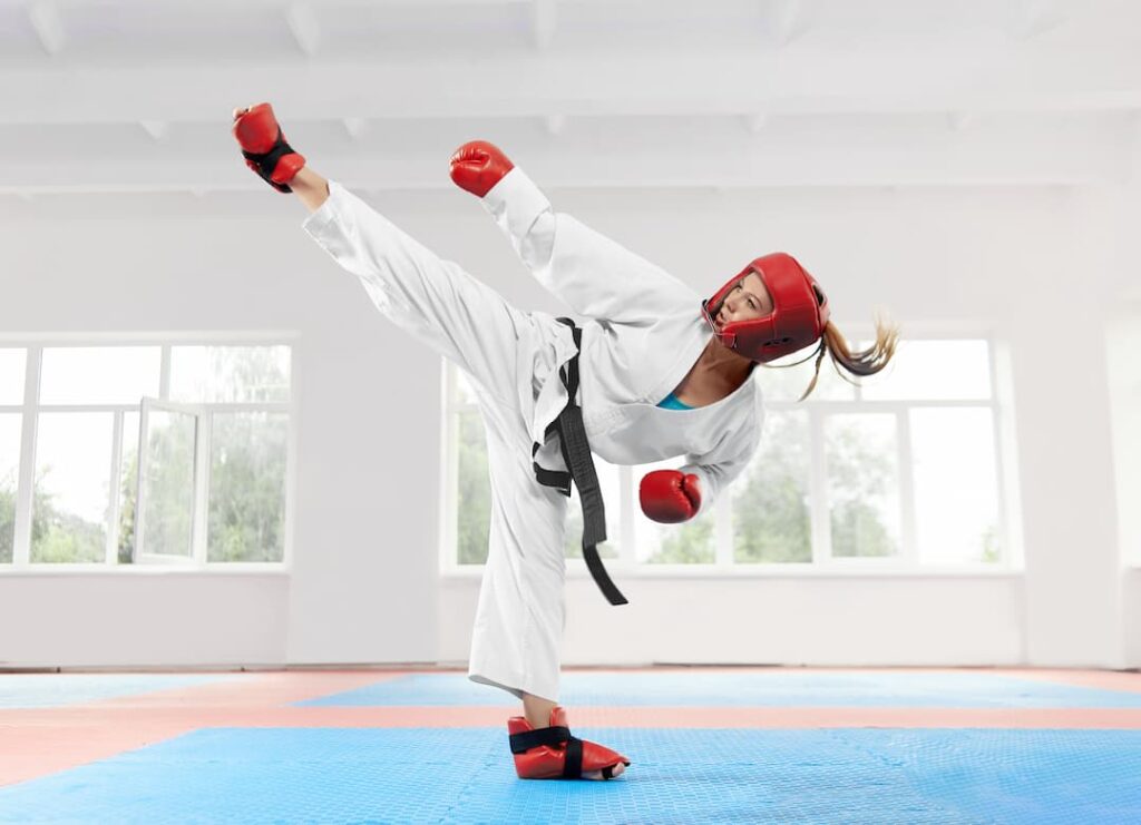 Femme en kimono pratiquant des arts martiaux avec des équipements de protection sur un tatami bleu dans un dojo lumineux.