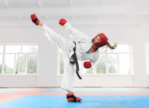Femme en kimono pratiquant des arts martiaux avec des équipements de protection sur un tatami bleu dans un dojo lumineux.