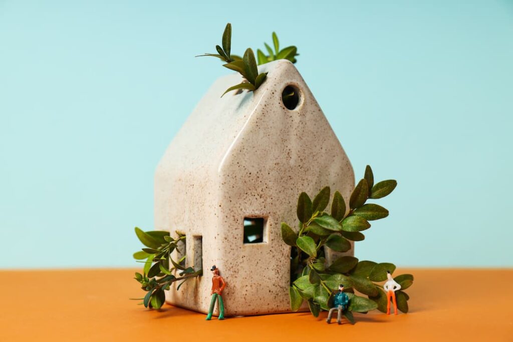 Petite maison écologique en céramique avec verdure luxuriante et figurines miniatures, symbolisant la construction durable et les matériaux comme les dalles écologiques pour sol.