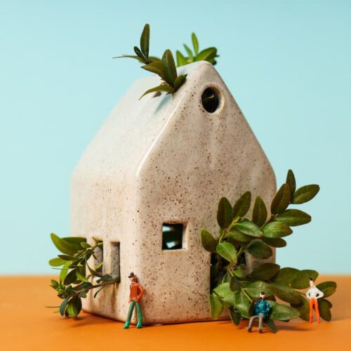 Petite maison écologique en céramique avec verdure luxuriante et figurines miniatures, symbolisant la construction durable et les matériaux comme les dalles écologiques pour sol.
