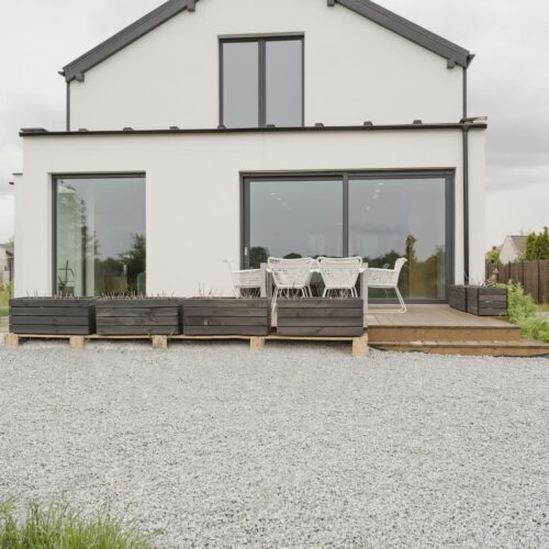 Maison moderne blanche avec terrasse en bois, mobilier extérieur et gravier. Idéal pour une terrasse sur sable ou des aménagements paysagers.