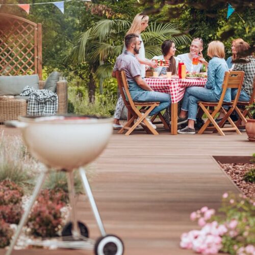 Des amis profitent d&#039;un barbecue et d&#039;un repas convivial sur une terrasse pas chère en bois, décorée de guirlandes, dans un jardin.