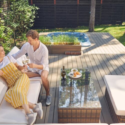Couple souriant se détendant sur une terrasse en bois clair avec jacuzzi, illustrant un aménagement réussi qui permet de comprendre et d&#039;éviter les erreurs lors de l&#039;installation d&#039;une terrasse composite.