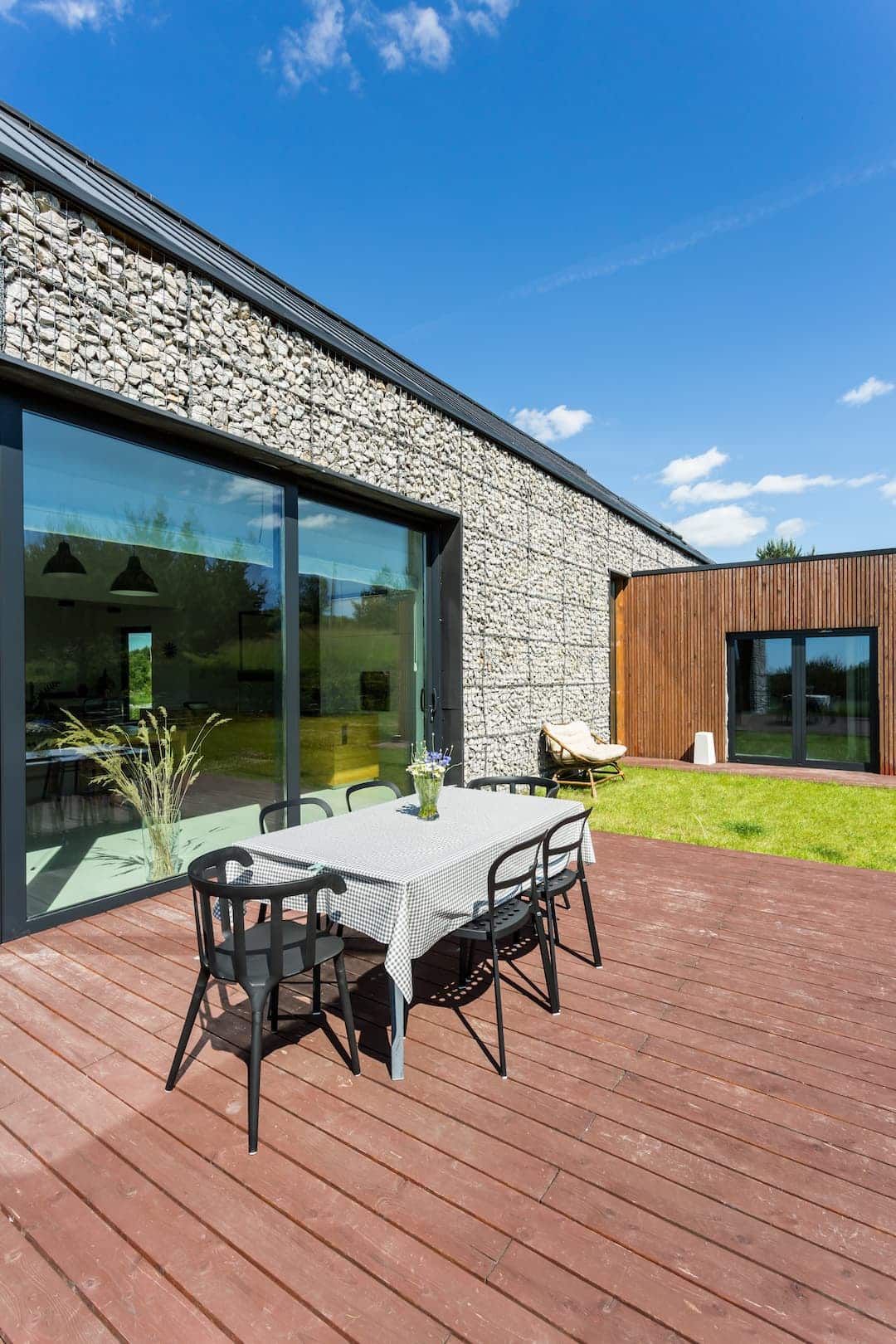 Maison moderne avec un meilleur bardage extérieur en gabion et bois, terrasse en bois, sous un ciel bleu. Salon de jardin avec table et chaises noires.